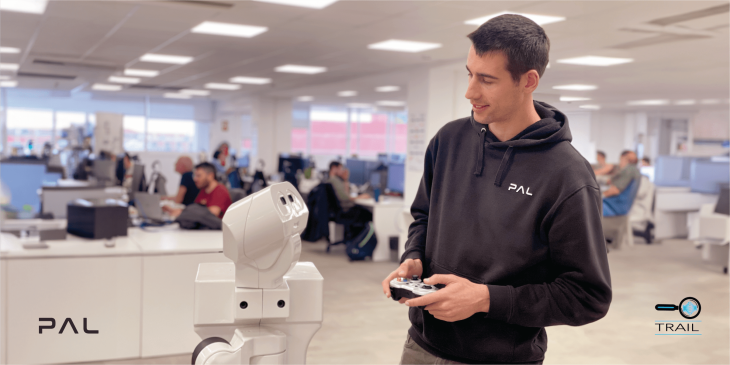 The image depicts Ferran Gebelli interacting with a TIAGo robot by PAL Robotics. The setting appears to be a busy office or research lab with several individuals at their workstations in the background. The focus is on Ferran and the robot, highlighting a moment of human-robot interaction. The logos for PAL Robotics and TRAIL are also visible, suggesting an innovative technology environment.
