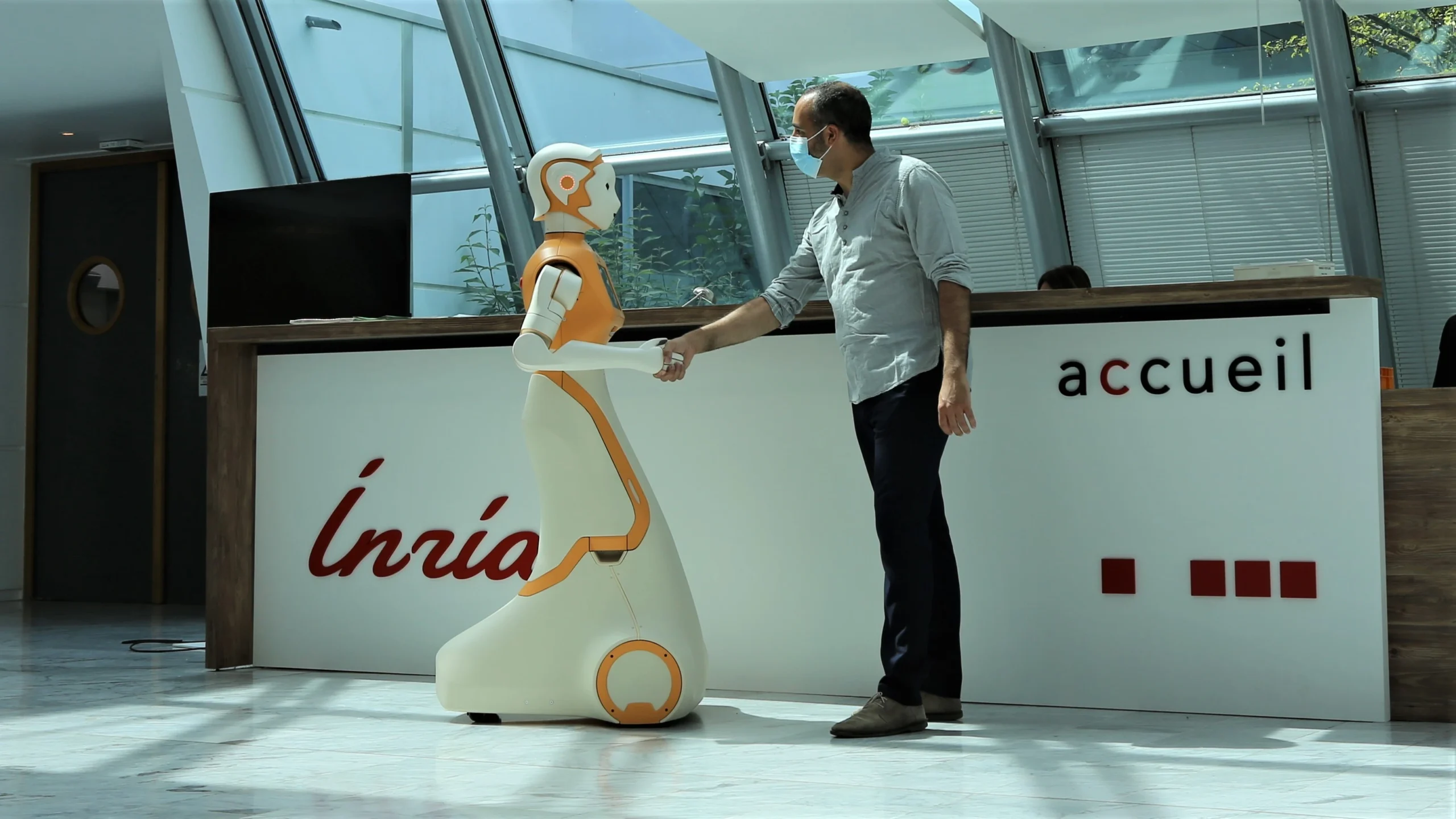 A man in a grey shirt and face mask is shaking hands with a humanoid robot named ARI in a modern lobby. The robot is white and orange, designed with a sleek, futuristic aesthetic. The background features a reception desk with the logo 'Inria' and a glass ceiling that lets in natural light.